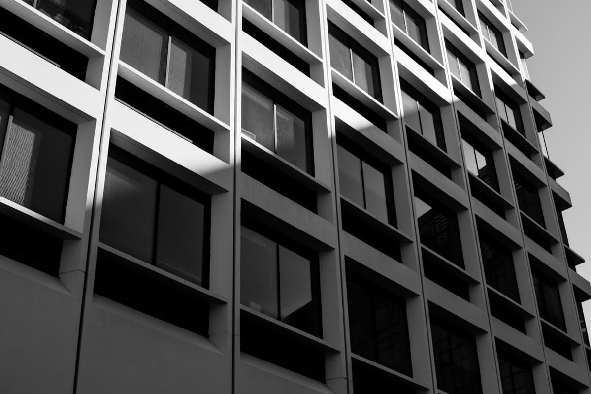 Modern facade with floor-to-ceiling window grid surrounded by lush greenery — each opening negotiates heat loss (U-value) against solar gain (SHGC) through the seasons